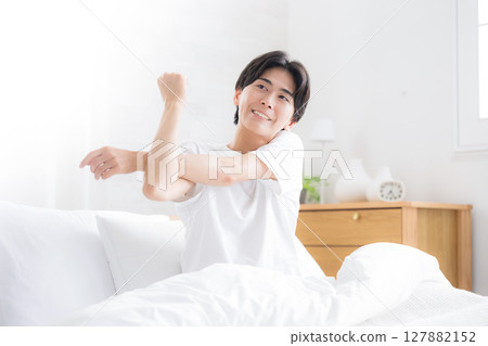 A young man waking up in bed bathed in the morning sun stretching his arms 127882152