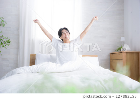 一位年輕男子在床上醒來,沐浴在清晨的陽光下,伸展雙臂,做出「萬歲」的姿勢 一位年輕男子在床上醒來,沐浴在清晨的陽光下,伸展雙臂,做出「萬歲」的姿勢 127882153