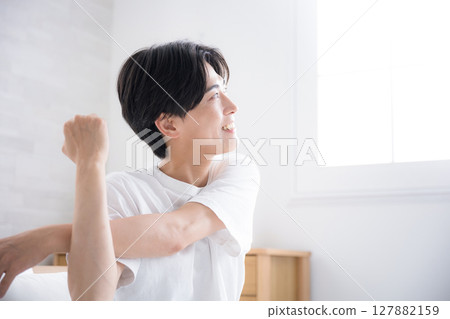 一位年輕男子在床上醒來，沐浴在早晨的陽光下，伸展雙臂，特寫 127882159
