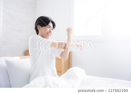 A young man waking up in bed bathed in the morning sun stretching his arms 127882160