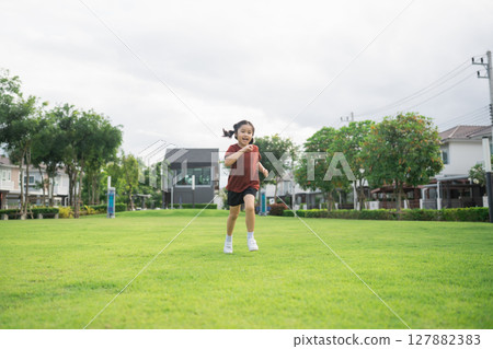 Joyful girl running in a green park during daytime, enjoying outdoor activity and vitality among trees and modern homes in the background 127882383