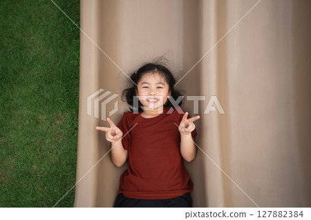 Joyful child playing on a slide outdoors, wearing a brown shirt, smiling while lying on a playground slide with a grassy background 127882384