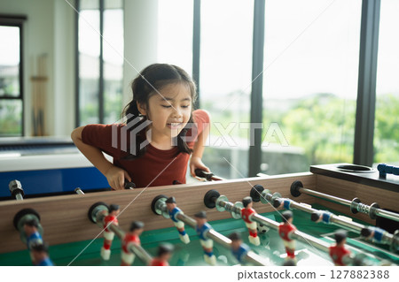 Joyful Girl Enjoys Foosball Game in Bright Room with Large Windows Overlooking Green Outdoors, Capturing a Moment of Playful Energy and Friendship 127882388