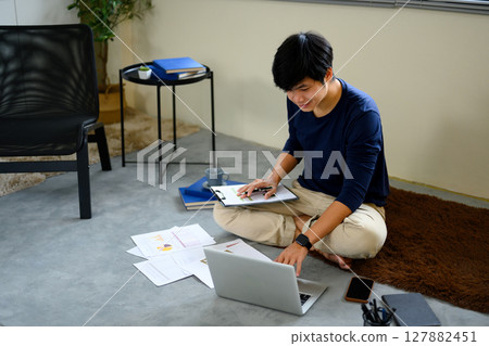Man managing personal finances at home, reviewing reports with laptop and documents on the floor Man managing personal finances at home, reviewing reports with laptop and documents on the floor 127882451