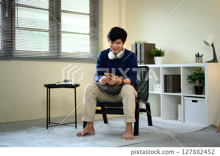 Relaxed asian man enjoying leisure time at home, using mobile phone for social media 127882452