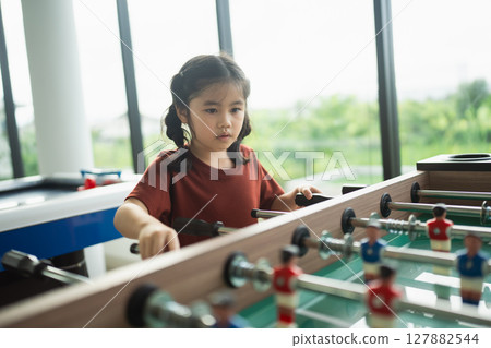 Young girl playing foosball in a bright room with large windows overlooking greenery, enjoying leisure time and developing skills through play 127882544