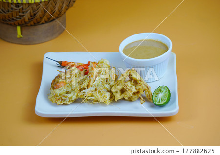 A plate of Bakwan or Indonesian fried snacks with a peanut sauce, lime, and chilies, set against a yellow and orange background with a traditional rattan container. A plate of Bakwan or Indonesian fried snacks with a peanut sauce, lime, and chilies, set against a yellow and orange background with a traditional rattan container. 127882625