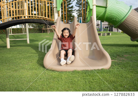 Excited Child Sliding Down Playground Slide with Arms Raised in Joy at Play Area on a Sunny Day Surrounded by Green Grass and Trees 127882633