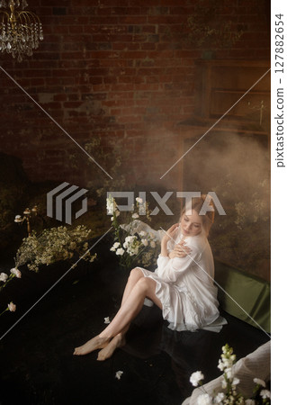 Young woman in white dress sitting in artificial pond with flowers and fog against brick wall 127882654