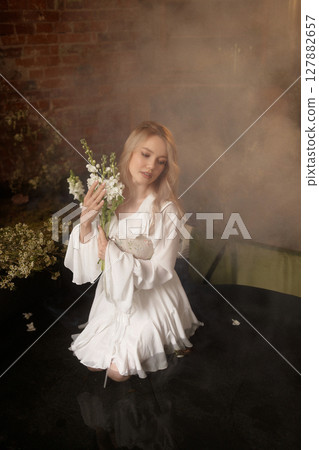 Young woman in white dress sitting in artificial pond with flowers and fog against brick wall 127882657