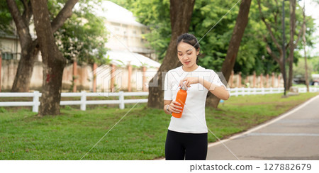 Active Lifestyle and Hydration. Young woman opening orange water bottle while walking in park. Active Lifestyle and Hydration. Young woman opening orange water bottle while walking in park. 127882679