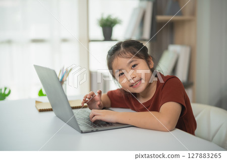 Smiling Asian girl working on laptop at home, engaging in creative activities, enjoying online learning, modern workspace, natural light, productive environment 127883265