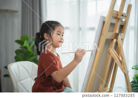 Young Girl Focused on Painting Creativity at Home with a Wooden Easel and Lush Green Plants in a Bright and Modern Interior Setting Young Girl Focused on Painting Creativity at Home with a Wooden Easel and Lush Green Plants in a Bright and Modern Interior Setting 127883285
