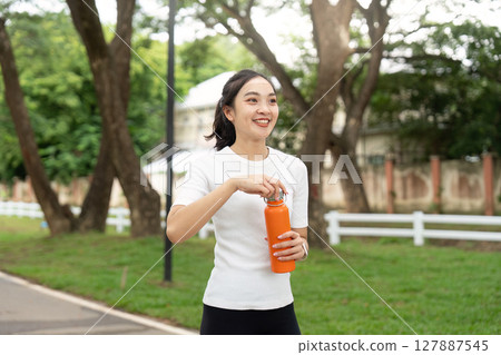 Active Lifestyle and Hydration. Young woman smiling while holding orange water bottle in park. 127887545
