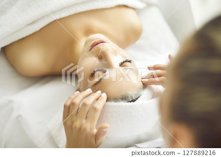 Beautician at spa is applying cosmetic sheet mask on face of beautiful young woman 127889216