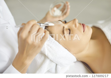 Female cosmetologist applying moisturizing mask with brush to a young woman in spa salon. 127889217