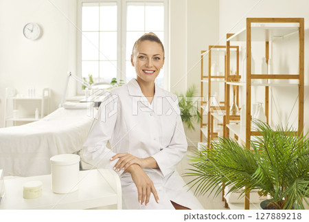 Portrait of a young smiling woman cosmetologist sitting in beauty spa salon and looking at camera. 127889218