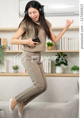 Joyful Music Experience at Home. Young woman dancing and singing with headphones in cozy living room. 127889825