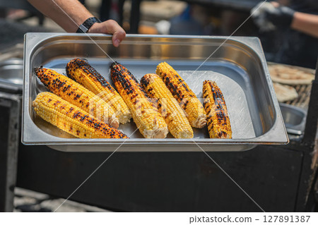 Close-up of grilled corn on cob. Fresh grilled corn on cob served in metal tray at street food festival. Concept of healthy snack, summer grilling, outdoor snack flavors Close-up of grilled corn on cob. Fresh grilled corn on cob served in metal tray at street food festival. Concept of healthy snack, summer grilling, outdoor snack flavors 127891387