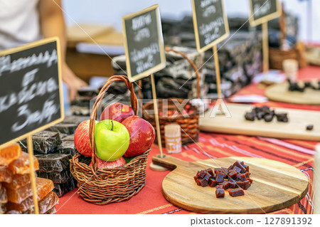 Colorful apples in wicker basket on market stall beside handmade fruit marmalade. Concept of fresh produce and artisanal natural fruit candy, marmalade presentation Colorful apples in wicker basket on market stall beside handmade fruit marmalade. Concept of fresh produce and artisanal natural fruit candy, marmalade presentation 127891392