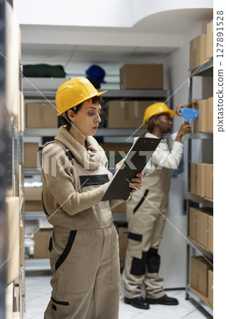 Woman warehouse worker ensures inventory management with boxes on racks, focusing on goods dispatch and quality control operations. Pallets stacked in the industrial storehouse. Woman warehouse worker ensures inventory management with boxes on racks, focusing on goods dispatch and quality control operations. Pallets stacked in the industrial storehouse. 127891528
