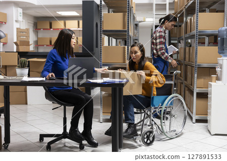 Asian woman in wheelchair working with her colleague to pack goods for the order shipment, doing product handling at the accessible packaging station. Local delivery and retail distribution. 127891533