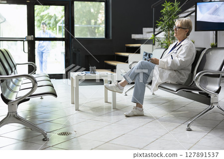 Senior doctor sitting in hospital lobby waiting for a patient to do scan interpretation, holding medical images on the bench. Healthcare worker in lab coat holding x rays preparing for checkup. 127891537