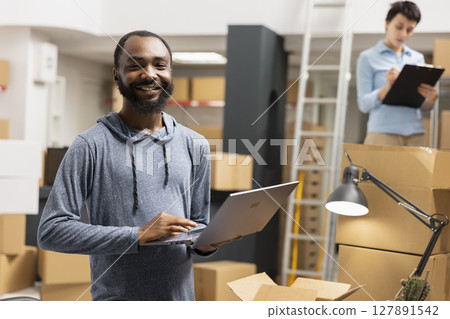 African american man overseeing product shipment and order logistics, working in small scale factory operations with taped boxes stacked on shelves. Handling goods and processing orders. African american man overseeing product shipment and order logistics, working in small scale factory operations with taped boxes stacked on shelves. Handling goods and processing orders. 127891542