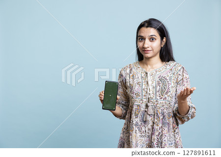 Portrait of indian woman doing marketing ad with green screen device, standing against blue background. Smiling young adult feeling positive on an advertisement in front of the camera. 127891611