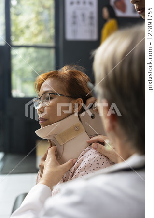 Medical staff fixing cervical collar on woman with signs of neck trauma, physiotherapy and recovery steps. Injured asian patient listens to prevention tips and treatment plan instructions. Medical staff fixing cervical collar on woman with signs of neck trauma, physiotherapy and recovery steps. Injured asian patient listens to prevention tips and treatment plan instructions. 127891755