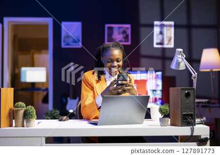 Happy woman at home having fun looking at social media reels done by online content creators. Smiling african american teenager relaxing by using mobile social network app at laptop desk Happy woman at home having fun looking at social media reels done by online content creators. Smiling african american teenager relaxing by using mobile social network app at laptop desk 127891773