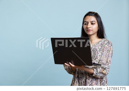 Indian young adult composing email or typing on her laptop in studio, solving her professional tasks on pc. Casual focused person browses web online standing against blue background. 127891775