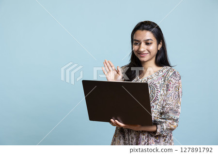 Confident upbeat girl sharing a story with best friend over web video call, standing against blue background with smartphone. Happy woman talking with mate online on teleconference. Confident upbeat girl sharing a story with best friend over web video call, standing against blue background with smartphone. Happy woman talking with mate online on teleconference. 127891792