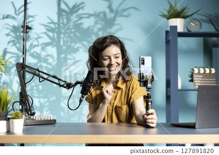 Cheerful internet star talking in front on cellphone attached to selfie stick in home studio, chatting with viewers. Happy woman using phone camera to record vlog for podcasting channel Cheerful internet star talking in front on cellphone attached to selfie stick in home studio, chatting with viewers. Happy woman using phone camera to record vlog for podcasting channel 127891820