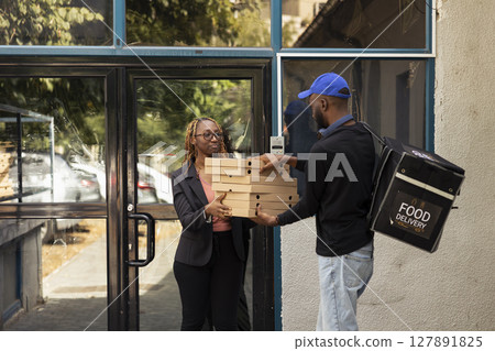 Black takeout courier handing a heavy pile of pizza boxes to a client, express takeout service. Male employee brings takeaway meal from a fast food restaurant in an urban setting. Black takeout courier handing a heavy pile of pizza boxes to a client, express takeout service. Male employee brings takeaway meal from a fast food restaurant in an urban setting. 127891825