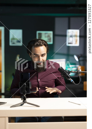 Man in studio endorsing smartphone from brand sponsoring video, talking in professional microphone. Tech specialist does influencer marketing by promoting mobile phone, recording with high quality mic 127891827