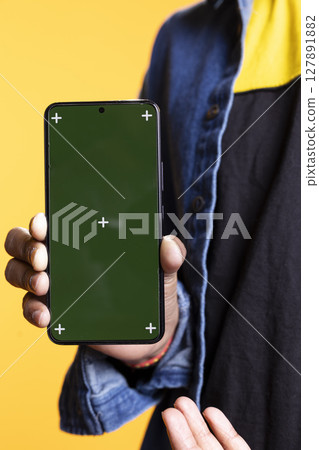 Indian content creator presenting green screen on mobile device, standing against orange background in the studio. Young male model wearing casual clothes and a man bun, internet channel. 127891882