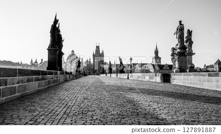 Sunrise casts a golden hue over Charles Bridge in Prague, revealing the cobblestone path and statues lining the bridge. The calm Vltava River reflects the city historic architecture. 127891887