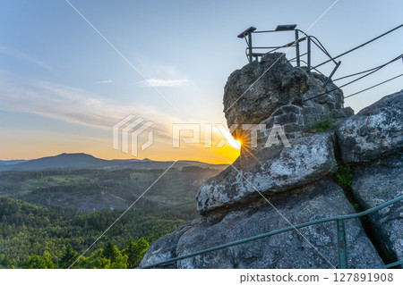 The sun sets behind Popov Cliffs, casting warm light over the sandstone rocks. Lush green forests stretch below, creating a stunning backdrop in Czechia's Lusatian Mountains during the evening. The sun sets behind Popov Cliffs, casting warm light over the sandstone rocks. Lush green forests stretch below, creating a stunning backdrop in Czechia's Lusatian Mountains during the evening. 127891908