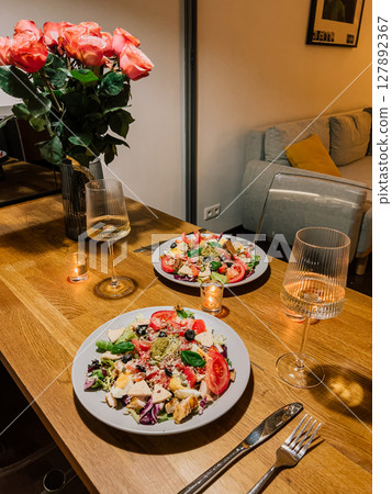 Intimate home dining experience with rose bouquet, candlelight, and healthy gourmet dishes on wooden table. Cozy romantic dinner setup for two with salad, candles, and wine in stylish home interior 127892367