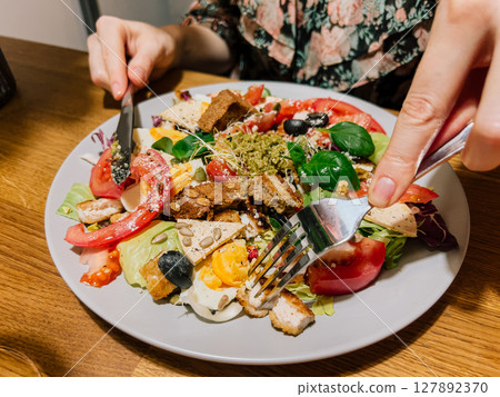 Woman eating homemade salad with fork and knife, eating moment with tomato and greens, eating plate with fresh flavors and natural ingredients. Bite into colorful food with eggs and pesto in cozy home 127892370
