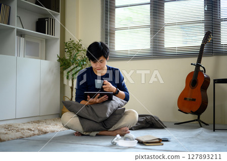 Young man sitting cross egged on the floor, drinking coffee and browsing tablet at home Young man sitting cross egged on the floor, drinking coffee and browsing tablet at home 127893211