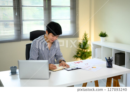 Businessman writing notes while working at desk with a laptop and documents Businessman writing notes while working at desk with a laptop and documents 127893240