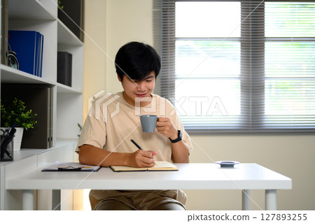 Young man enjoying morning coffee while writing notes in a notebook at home office Young man enjoying morning coffee while writing notes in a notebook at home office 127893255