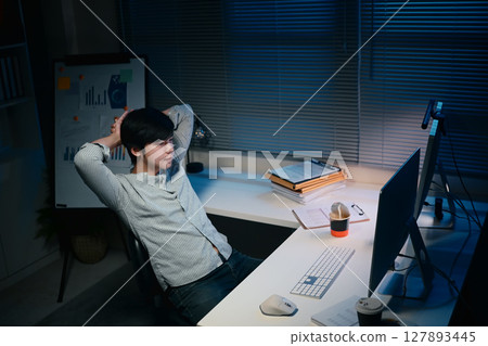 Tired male employee sits in a dark office, working overtime under dim lighting 127893445
