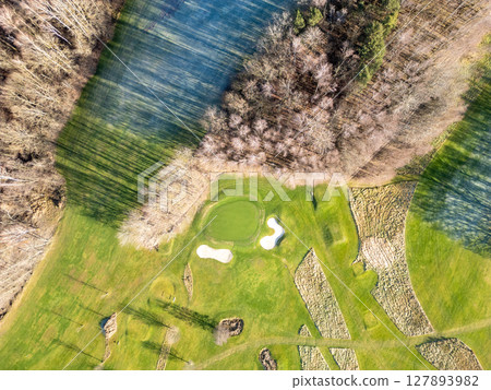 Elevated perspective of a well-maintained golf course surrounded by trees. The lush green fairways and sand traps are visible under clear skies. Ideal for golfing enthusiasts. 127893982