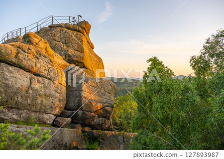 The warm glow of sunset illuminates the sandstone Popov Cliffs in Czechia. Lush greenery surrounds the rocky formation, showcasing the beauty of nature in the evening light. 127893987