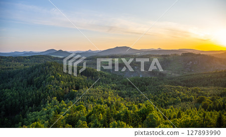 A breathtaking sunset casts golden hues over the Lusatian Mountains, viewed from the heights of Popov Cliffs. Dense forests stretch across the landscape, creating a serene evening atmosphere. 127893990