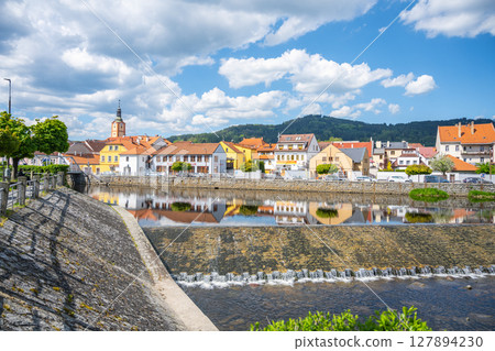 The tranquil waters of the Otava River reflect colorful buildings in Susice, Czechia. Visitors enjoy a sunny day with a backdrop of green hills and blue skies. 127894230