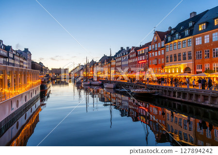 As dusk falls over Nyhavn, the waterfront glows with warm lights reflecting on the calm canal. People enjoy the vibrant atmosphere, dining and exploring the historic district. 127894242